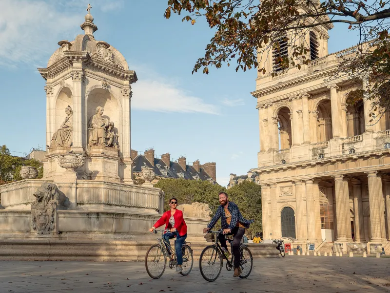 Deux cyclistes place Saint-Sulpice à Paris, devant la fontaine et l'église, au départ de la Véloscénie Paris-Mont-Saint-Michel.