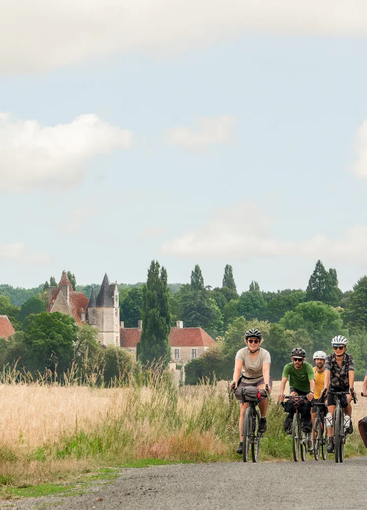 Groupe de cyclotouristes pédalant entre champs de blé et manoir de la Vôve, itinéraire Véloscénie dans le Perche